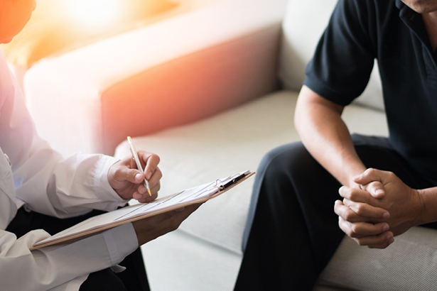 Mental health therapist writing notes on a clipboard next to a patient with clasped hands sitting on a couch